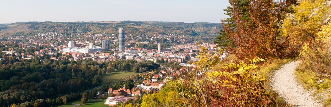 Ein sonniger, etwas höher gelegener, von rechts ins Bild kommender Wanderweg gibt  den Blick auf das Stadtzentrum von Jena frei